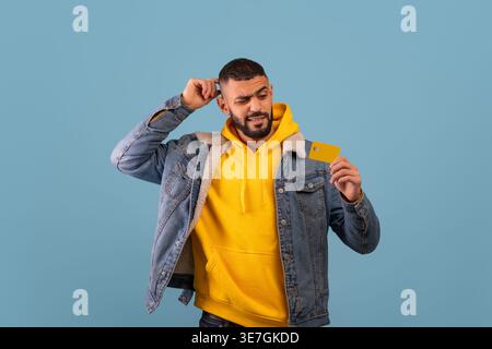 Middle Eastern man with beard wearing yellow hoodie and denim jacket holding credit card and touching head standing against blue background in studio Stock Photo