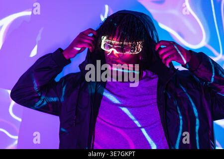 Black male gamer wearing red led augmented reality glasses adjusting hoodie and looking away while standing in dark room with colorful neon lights Stock Photo