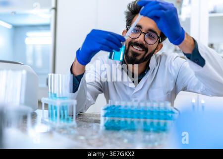 Smiling Indian male scientist wearing lab coat and latex gloves sitting at table and testing chemical sample in flask in laboratory Stock Photo