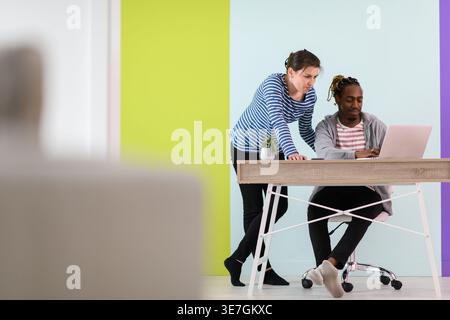 Black businessman with braids sitting at table working on laptop and having discussion with caucasian female colleague standing beside him in the o... Stock Photo
