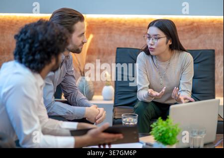Asian business woman wearing eyeglasses sitting on single couch looking at male coworkers while discussing ideas during meeting in office Stock Photo