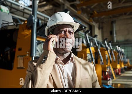 Black male engineer wearing safety helmet having phone call standing in warehouse during daytime Stock Photo