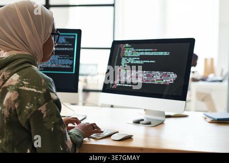 Focused Mixed-race Muslim female software developer decoding data working on computer with multiple screens sitting at table in office Stock Photo