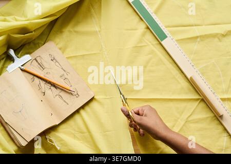 Top view of Black female fashion designer cutting yellow fabric using scissors on table with ruler, pencil, and fashion sketchbook in tailoring wor... Stock Photo