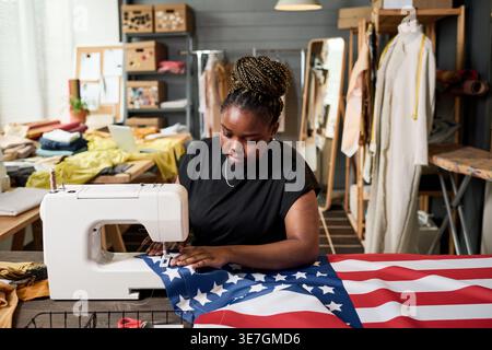 Serious Black woman sewing American flag using electric sewing machine sitting in tailor shop during daytime Stock Photo