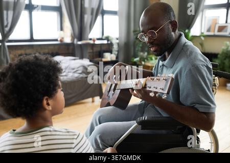 Smiling disabled Black father in glasses sitting in wheelchair playing guitar together with son at home Stock Photo
