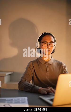 Asian man wearing headphones working on laptop sitting at desk in workplace at night Stock Photo