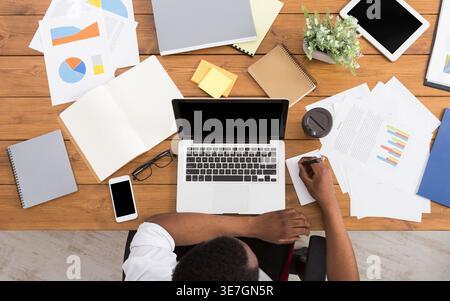 Black businessman taking notes sitting at wooden desk with laptop and various objects in office Stock Photo