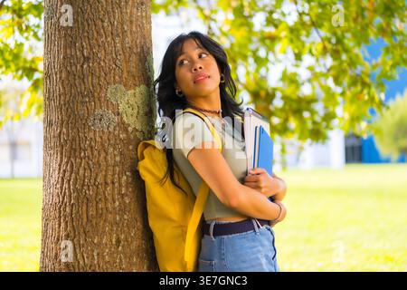 Asian female student with backpack holding books looking away leaning against tree trunk in university during daytime Stock Photo