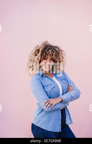 Smiling Hispanic woman with curly afro standing with crossed arms against pink background Stock Photo
