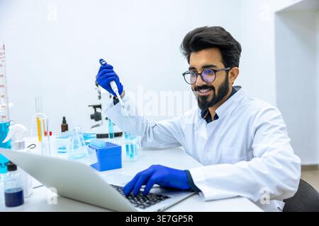 Smiling Indian male scientist wearing lab coat and latex gloves holding glass dropper with reagents and making notes on laptop while sitting at his... Stock Photo