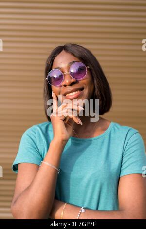 Smiling Black female in sunglasses touching face while standing against brown wall Stock Photo