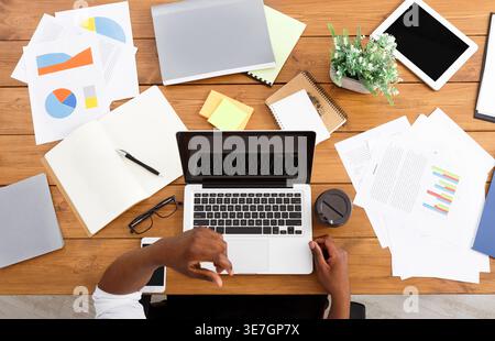Black businessman sitting at wooden desk with laptop and various objects in office Stock Photo