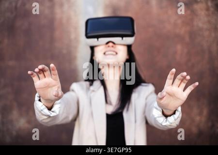 Smiling Asian woman reaching hands forward wearing VR headset standing against shabby wall during daytime Stock Photo