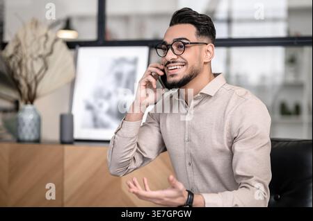 Smiling Arab man wearing eyeglasses having phone conversation standing at home Stock Photo