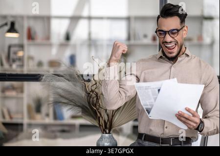 Cheerful Arab man wearing eyeglasses raising arm with closed fist while reading paper standing at home Stock Photo