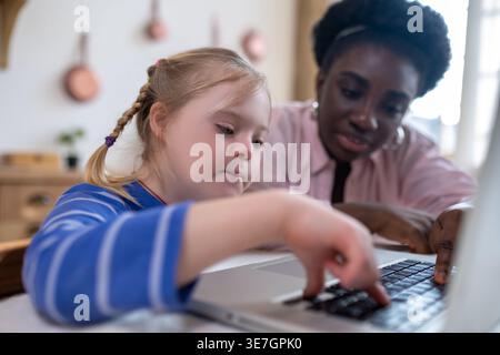 Black female tutor watching caucasian girl with down syndrome using laptop sitting at table at home Stock Photo
