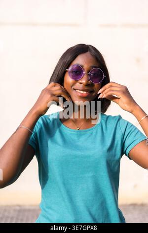 Smiling Black female in sunglasses touching face while standing against white wall Stock Photo