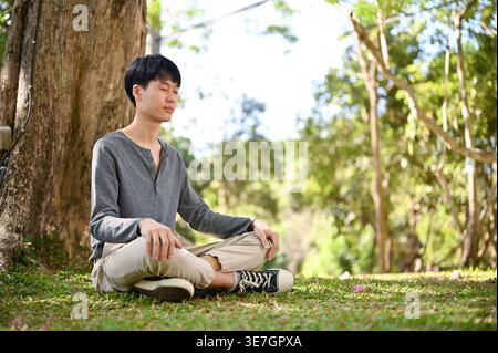 Asian male sitting with closed eyes and crossed legs on grass in park on sunny day Stock Photo