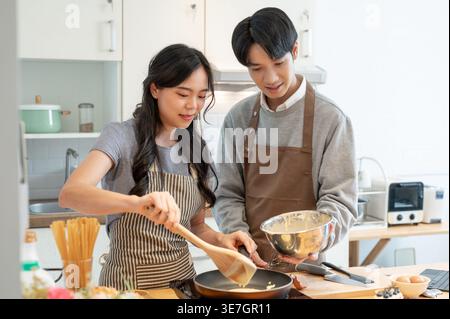 Asian couple wearing aprons having fun making pancakes together in kitchen at home Stock Photo