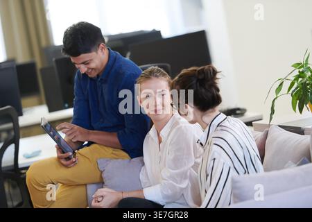 Middle Eastern man using tablet sitting on sofa beside caucasian female coworkers talking in office during break Stock Photo