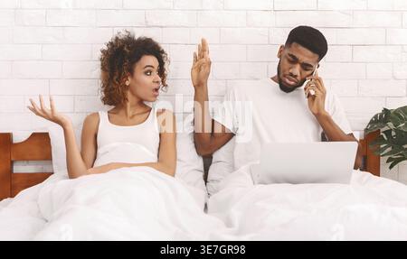 Angry Black woman with afro hair talking to husband having phone call while sitting on bed at home Stock Photo