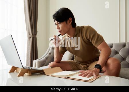 Asian male entrepreneur using laptop and taking notes in planner sitting on the couch at home Stock Photo