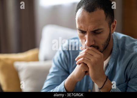 Sad Black man leaning mouth on clasped hands sitting on sofa in living room at home Stock Photo