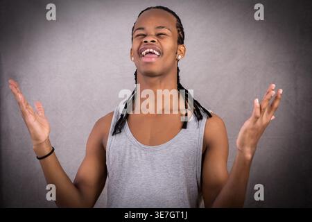 Young Black man with braided hair standing against gray background lifting both hands Stock Photo