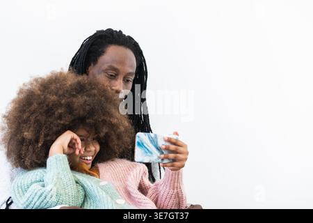Black father with locks hugging Mixed race daughter with afro while sitting and watching video on smartphone against white background in studio Stock Photo