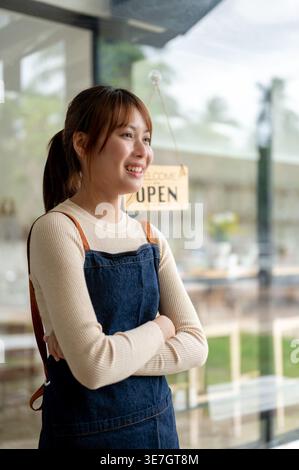Smiling Asian woman with crossed arms looking away standing near entrance door in coffee shop Stock Photo