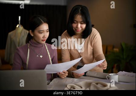 Asian female dressmaker using laptop while working with her assistant in tailor shop Stock Photo