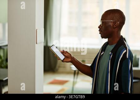 Black man using smartphone while connecting to smart home system standing indoors Stock Photo