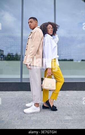 African American couple in stylish outfits standing leaning on each other's back against glass wall on street during daytime Stock Photo
