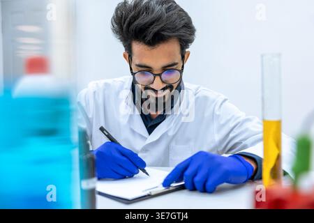 Smiling Indian male scientist wearing lab coat and latex gloves sitting at desk writing notes in laboratory Stock Photo