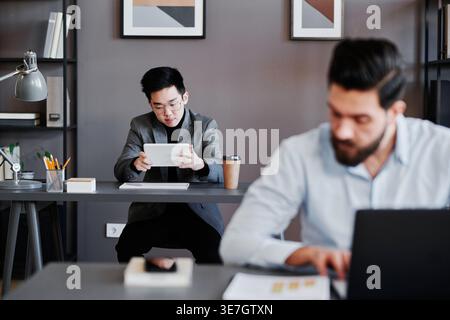 Serious young Asian businessman working on tablet sitting at table near male colleague in office Stock Photo