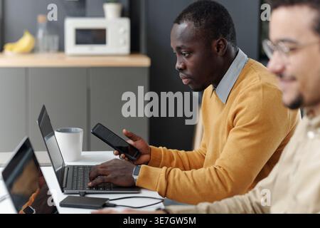 Black man holding smartphone working on laptop sitting at table with Middle Eastern colleague in office Stock Photo