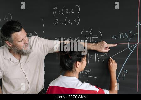 Asian female student solving math problem on blackboard with middle-aged caucasian male teacher helping her while standing in the classroom Stock Photo