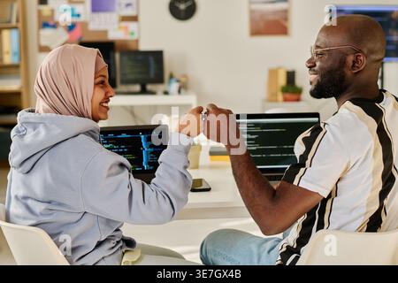 Black male and Mixed race Muslim female programmers doing fists bump sitting at desk with laptops in office Stock Photo
