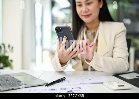 Smiling Asian business woman using smLifestylephone while sitting at table with document reports and laptop in office Stock Photo