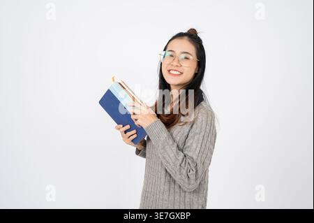 Smiling Asian female wearing eyeglasses holding books standing against gray background in studio Stock Photo