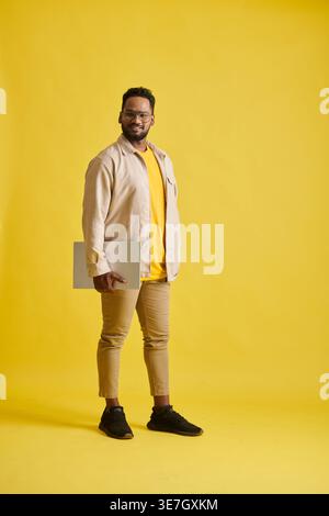 Smiling Asian man with eyeglasses carrying laptop standing against yellow background in studio Stock Photo