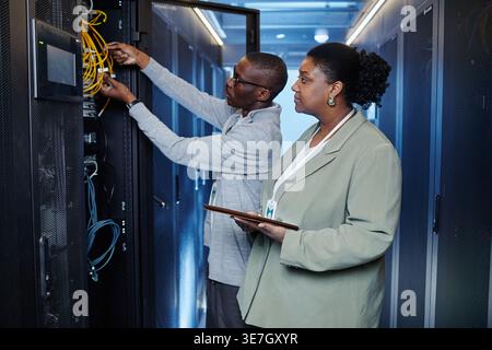 Black male IT engineer with eyeglasses connecting cables near Black female colleague holding tablet standing in server room in data center Stock Photo