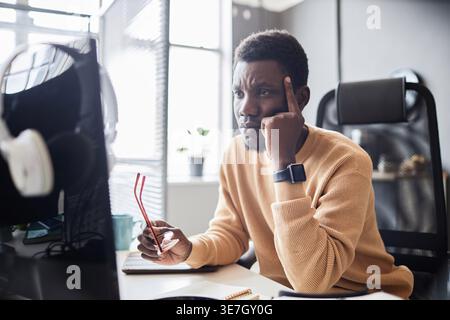 Black male programmer pointing finger on his temple holding eyeglasses looking at computer monitor sitting at table in office Stock Photo