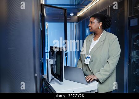Serious Black female IT engineer looking at cables standing at table with laptop in server room in data center Stock Photo