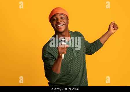 Black man in beanie singing using microphone while standing against yellow background in studio Stock Photo