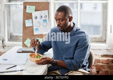 Black man holding a bowl of salad sitting at the table in office Stock Photo