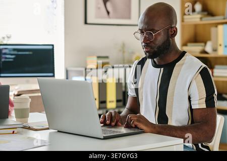 Black male programmer wearing eyeglasses typing codes on laptop while sitting at desk in office Stock Photo