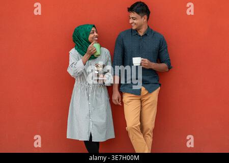 Black woman wearing hijab and Middle Eastern man looking at each other while having coffee standing against orange wall in street Stock Photo