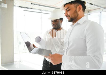 Diverse group of male engineers wearing hard hats holding blueprint and laptop discussing project during work on construction site Stock Photo